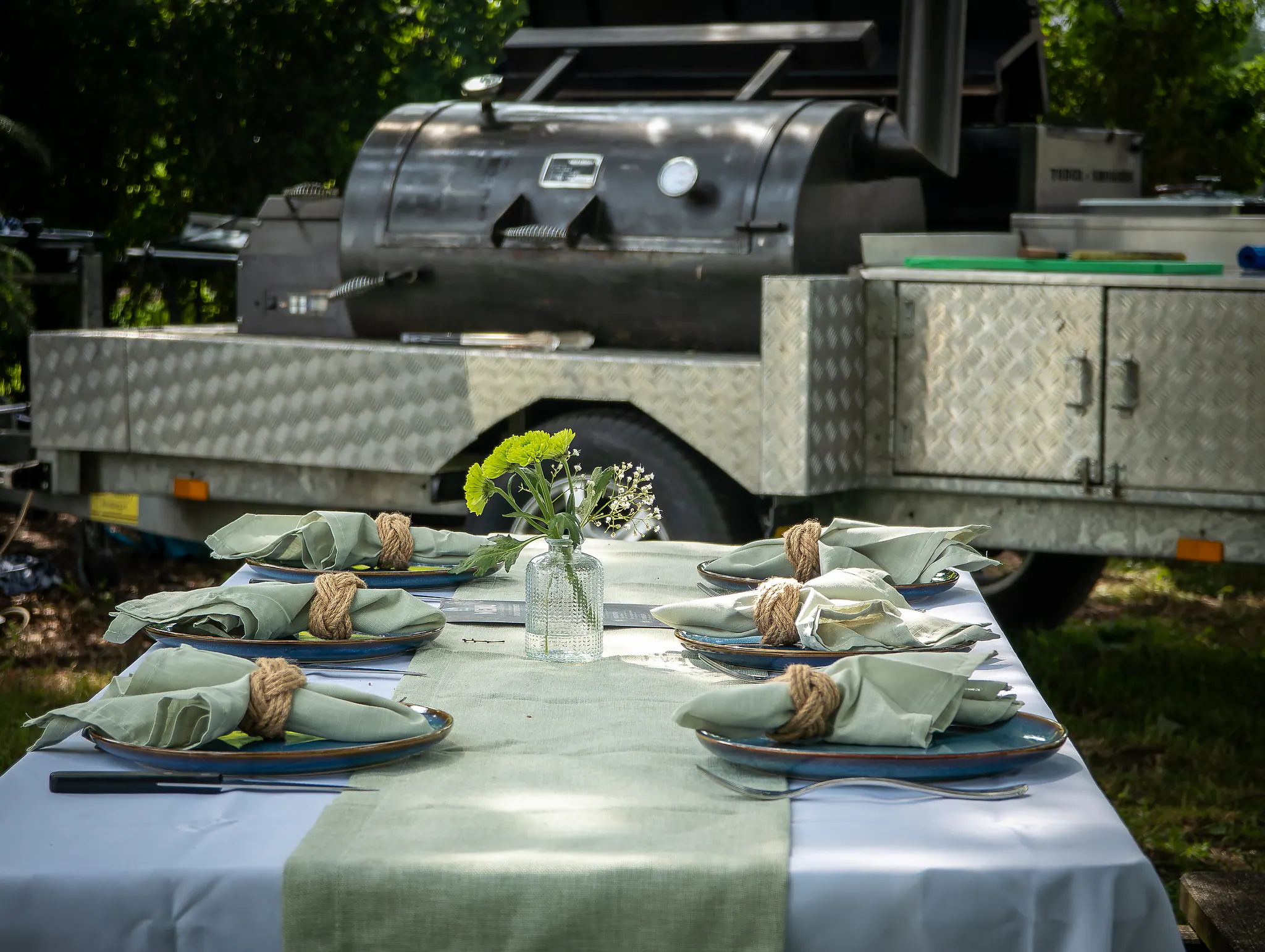 Een gedekte tafel buiten met linnen servetten, de smoker op de trailer erachter.